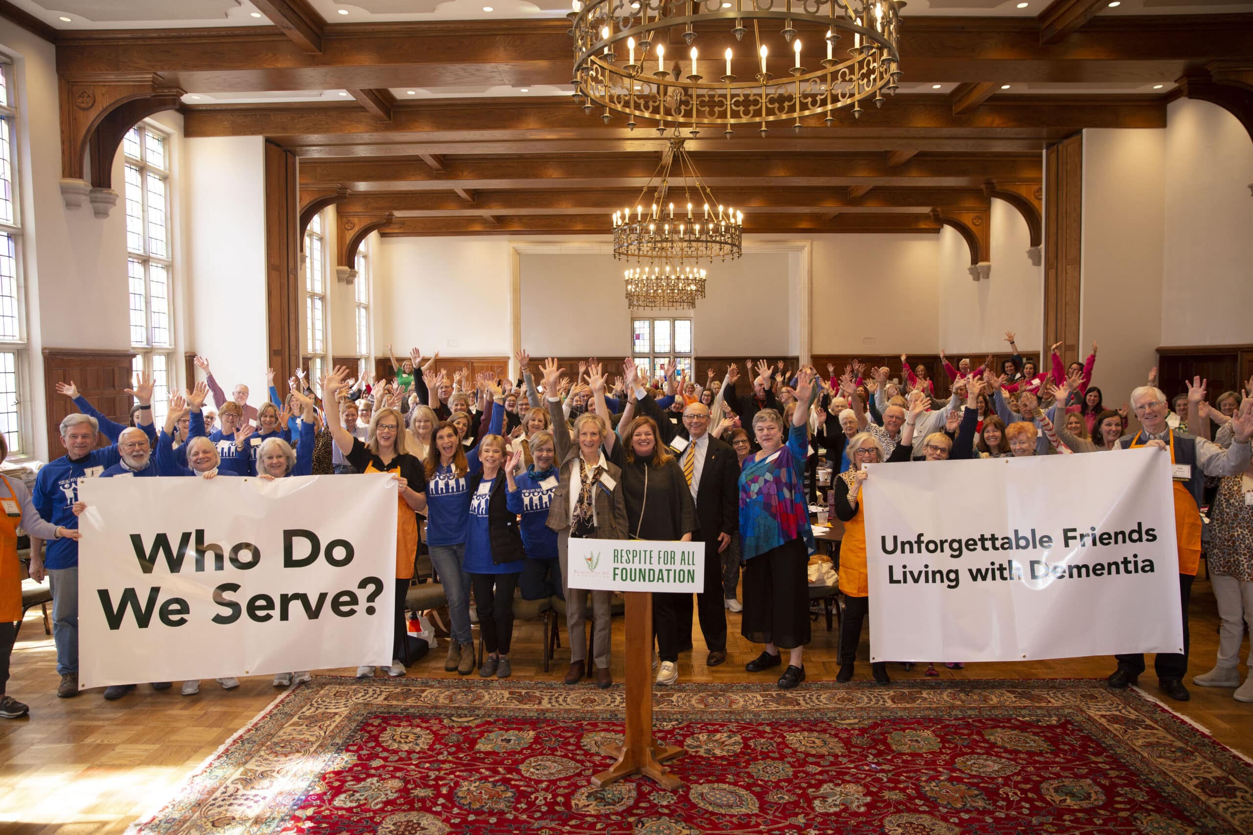 Large group indoors raising hands with signs “Who Do We Serve?” and “Unforgettable Friends Living with Dementia,” supporting Respite for All Foundation.