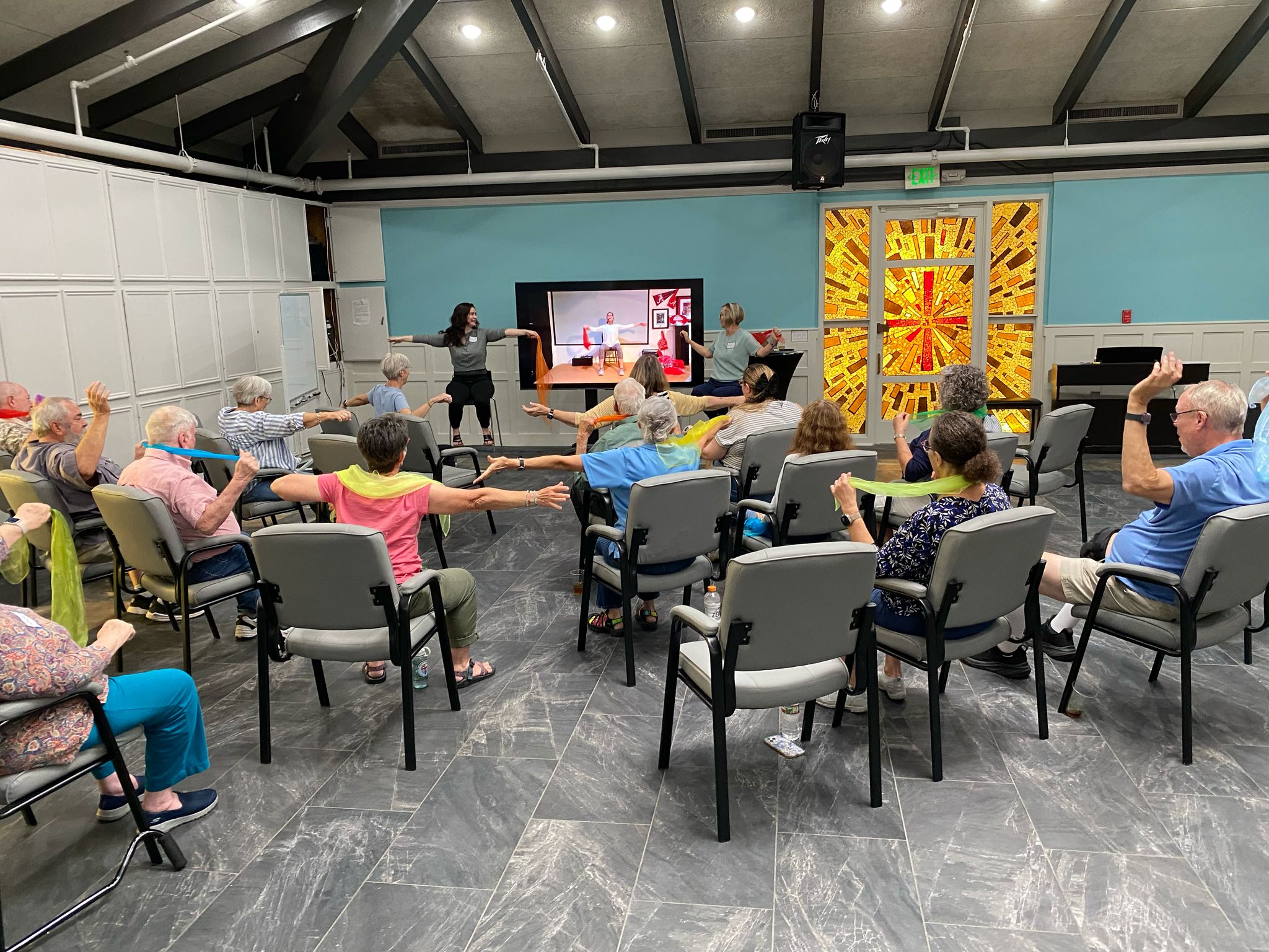 Participants in Houston RFA community seated in a circle, following two instructors leading a stretching exercise with scarves in a bright room.